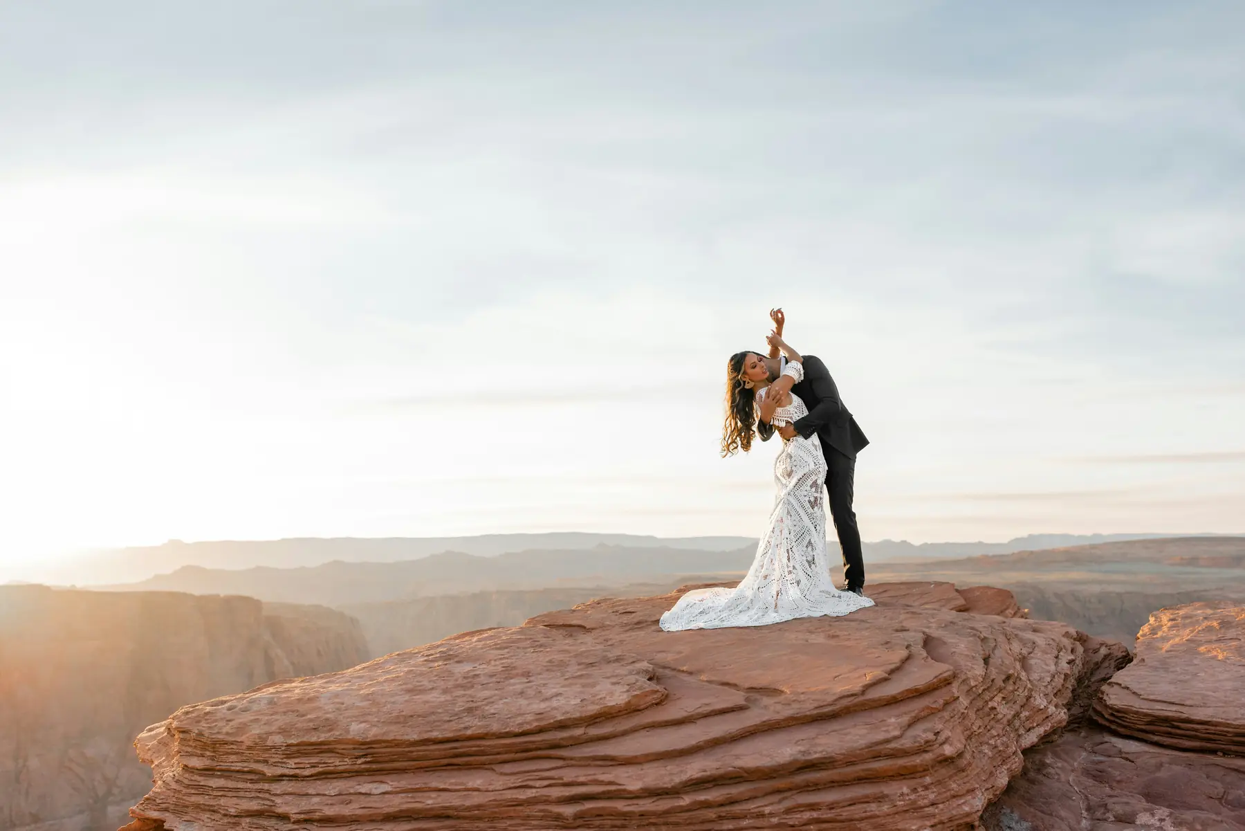A bride and groom dip into a kiss on a canyon rim at golden hour, her gown trailing over the red rock