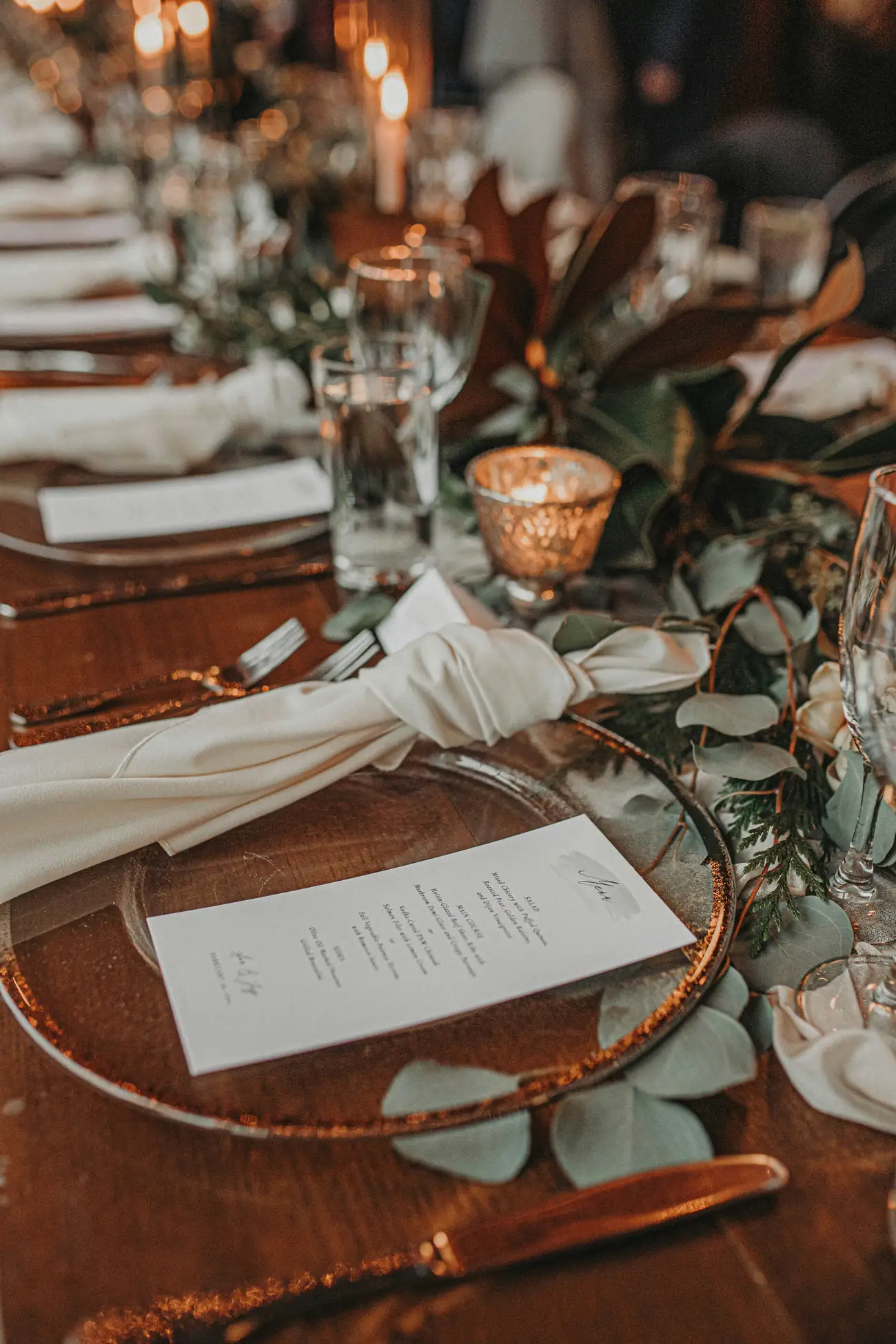 A candlelit table set with hand-lettered menus, knotted linen, eucalyptus and gold-rimmed glass